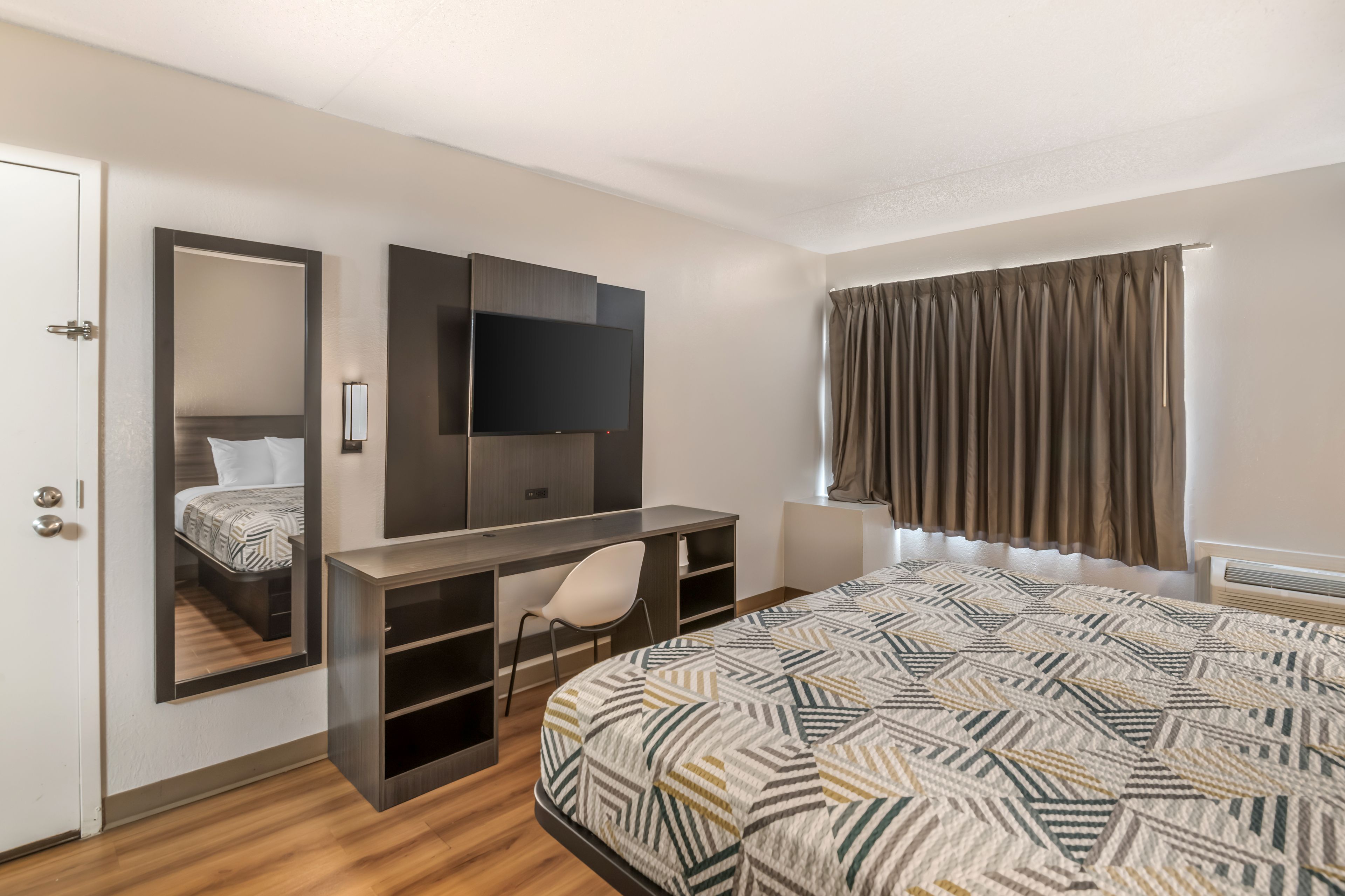 A hotel room with a bed, a desk with a chair, a flat-screen TV, a mirror, and a window with curtains.