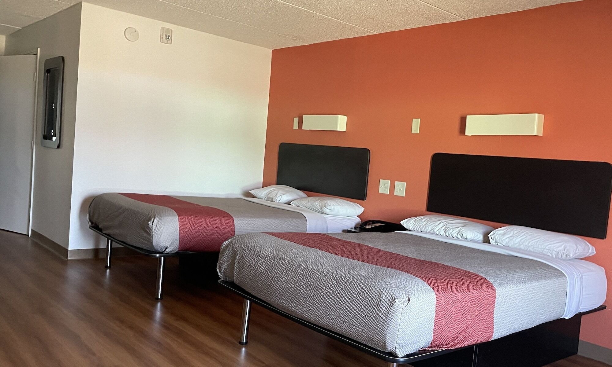 A hotel room with two beds, a wooden floor, and a plain wall with a thermostat.