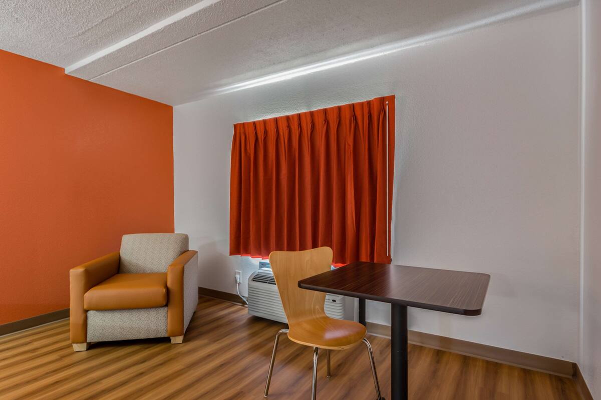 Ein Zimmer mit Holzboden, einem braunen Ledersessel, einem Holztisch und einem Fenster mit orangefarbenen Vorhängen.
