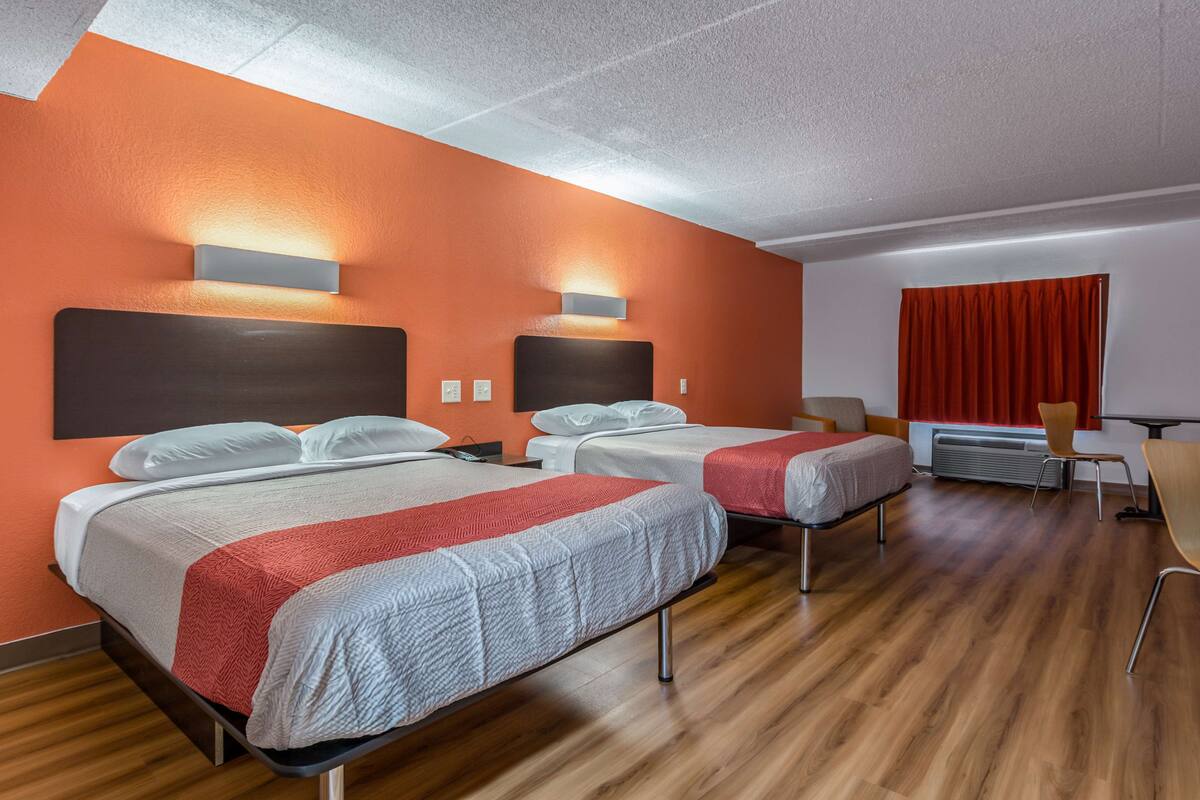 Ein Hotelzimmer mit zwei Betten, einem Schreibtisch, einem Stuhl und einem Fenster mit roten Vorhängen.