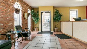 A reception area with a brick wall, a reception desk, two potted plants, a black leather chair, and a small table with a clock.