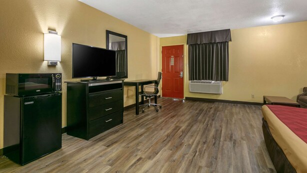 A hotel room with a flat-screen TV, a mini-fridge, a desk with a chair, a bed, and a red door.