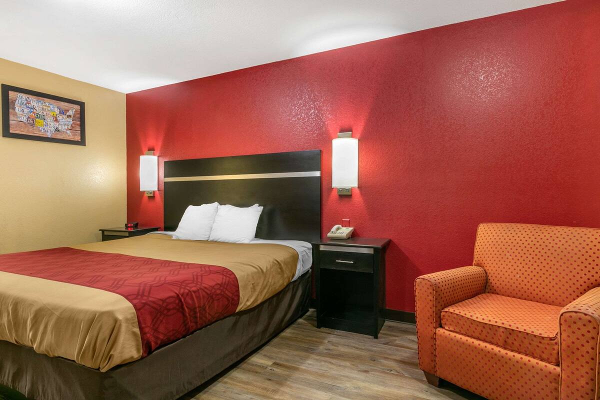 A hotel room with a bed, a nightstand, a chair, and a red wall.