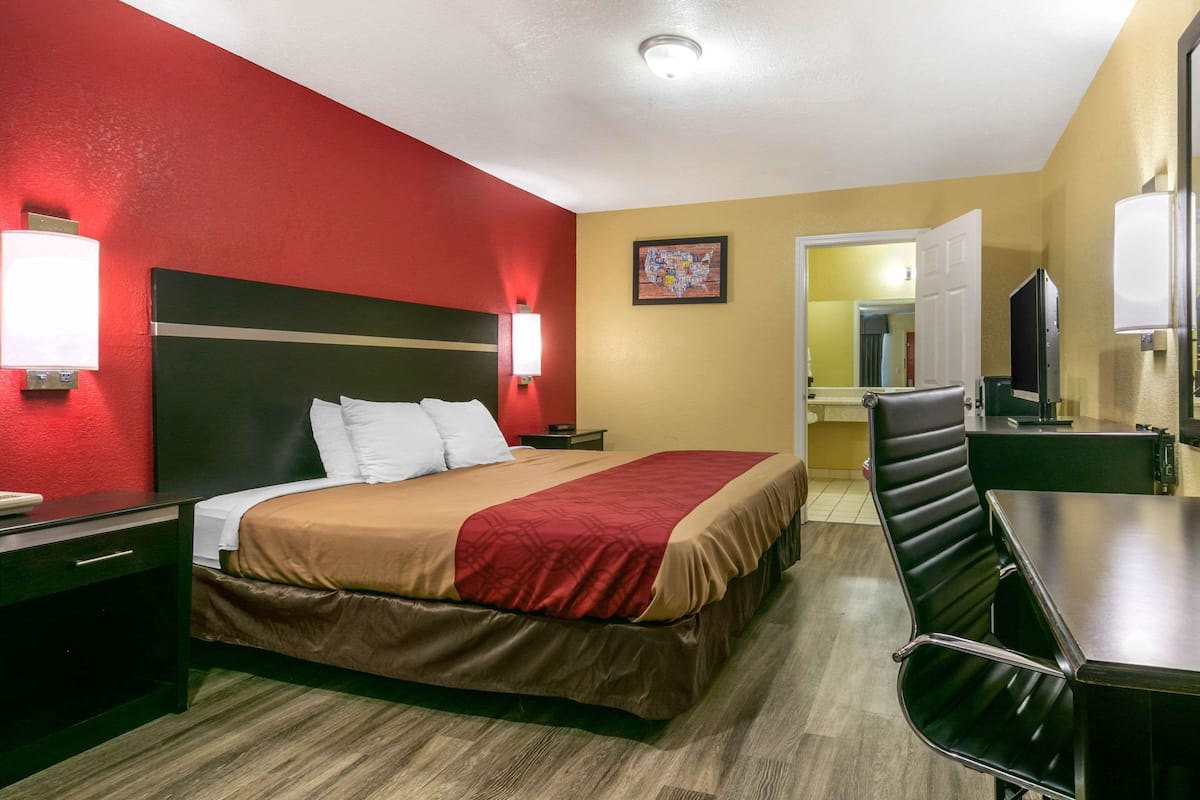 A hotel room with a bed, bedside table, lamp, and a desk with a chair.