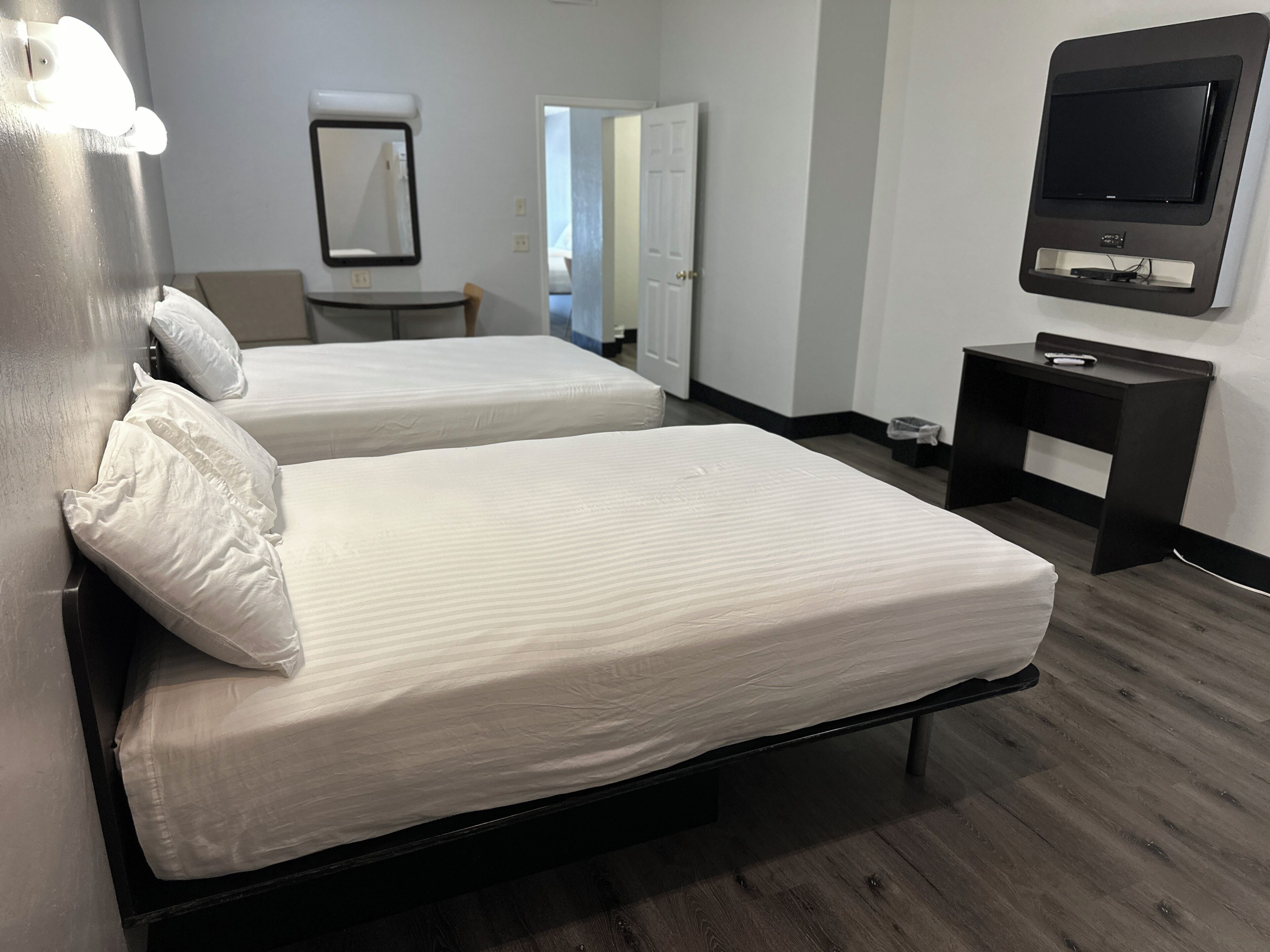 A hotel room with two single beds, a bedside table with a lamp, a TV mounted on the wall, and a door leading to another room.