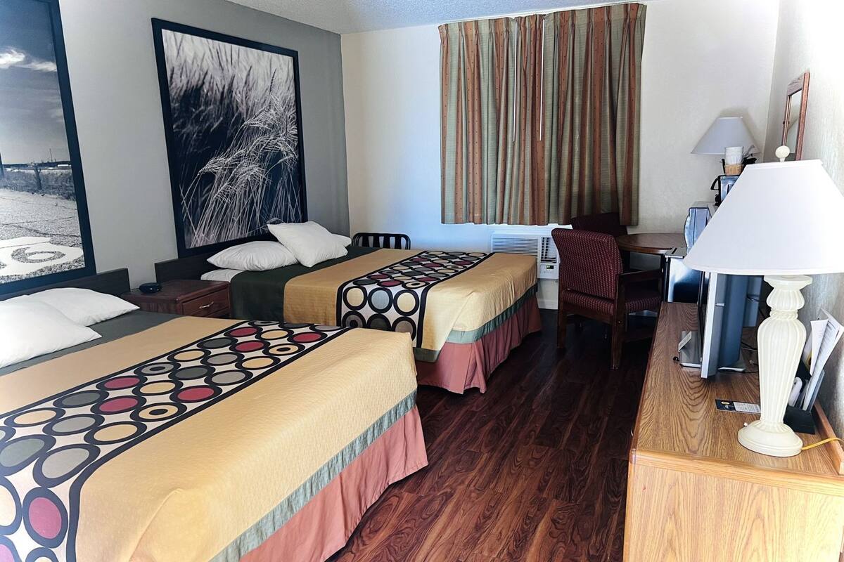 A hotel room with two beds, a desk, a chair, a lamp, and a large window with curtains.