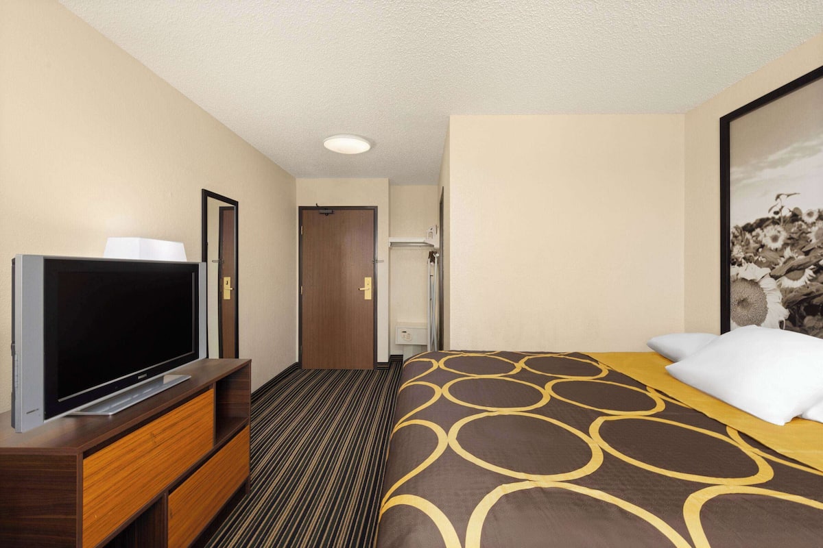 A hotel room with a flat-screen TV on a wooden TV stand, a bed with a patterned bedspread, and a framed picture on the wall.
