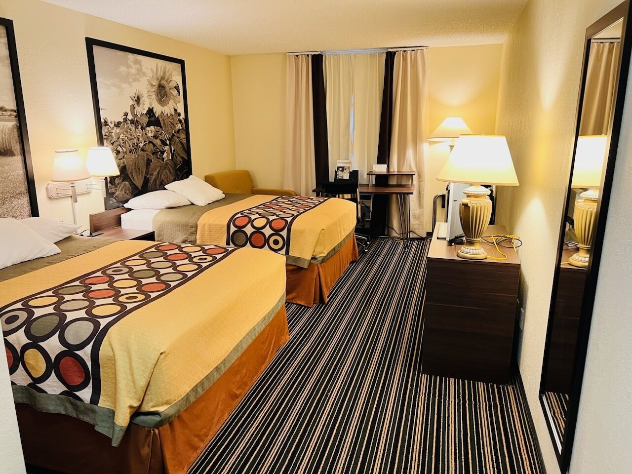 A hotel room with two beds, a desk, a chair, a lamp, and a mirror.