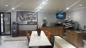 A hotel dining area with a long table, chairs, an ATM, a large Texas sign, a television, and a kitchen area with a coffee machine.