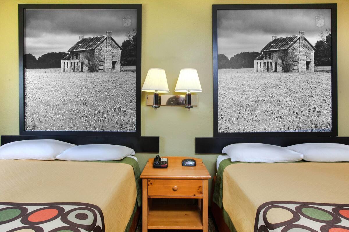 Habitación de hotel con dos camas, una mesita de noche con lámparas y dos fotografías blanco y negro de una casa en un campo en las paredes.