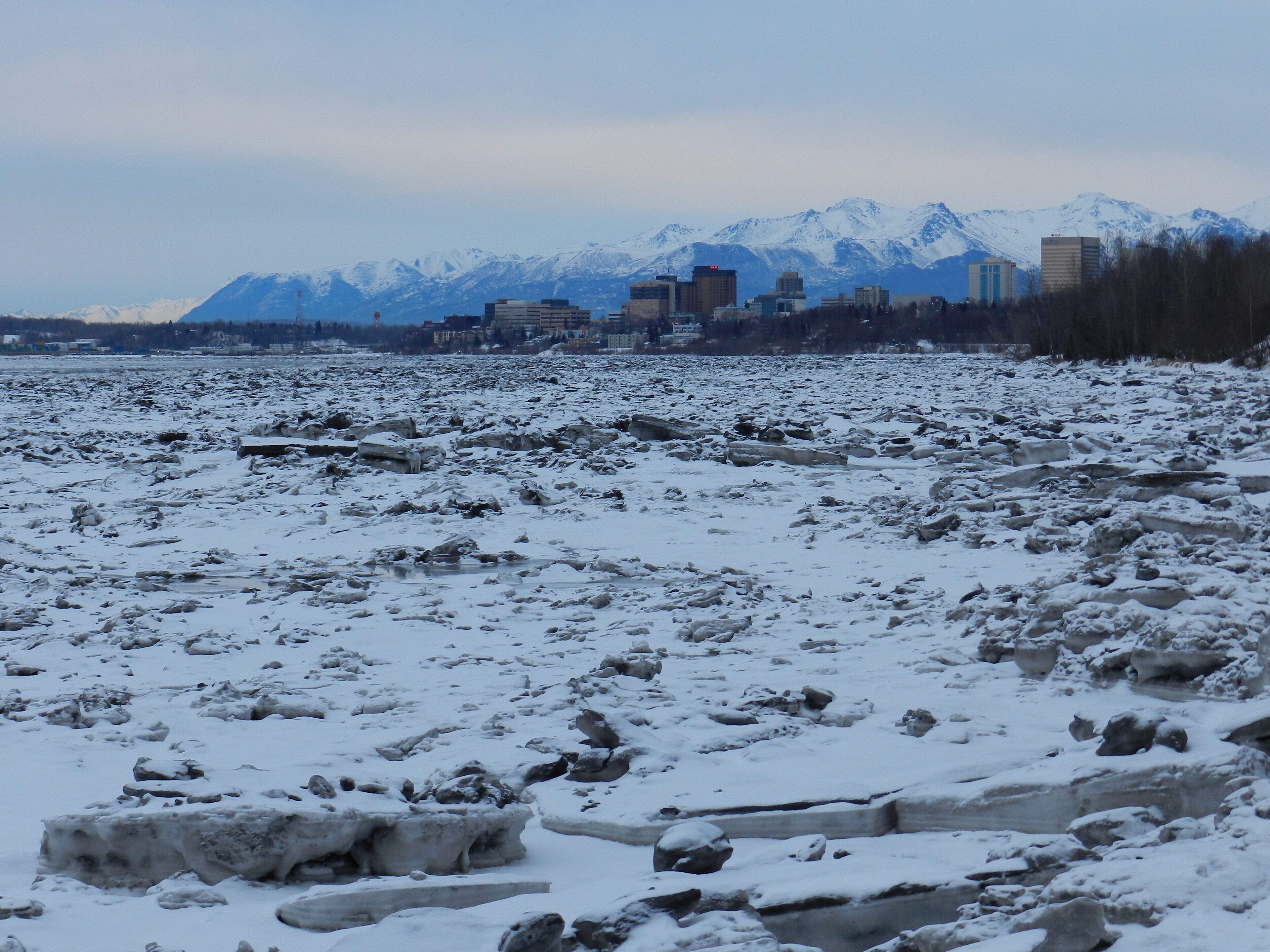 A snowy landscape with a river, ice formations, and a cityscape with mountains in the background.