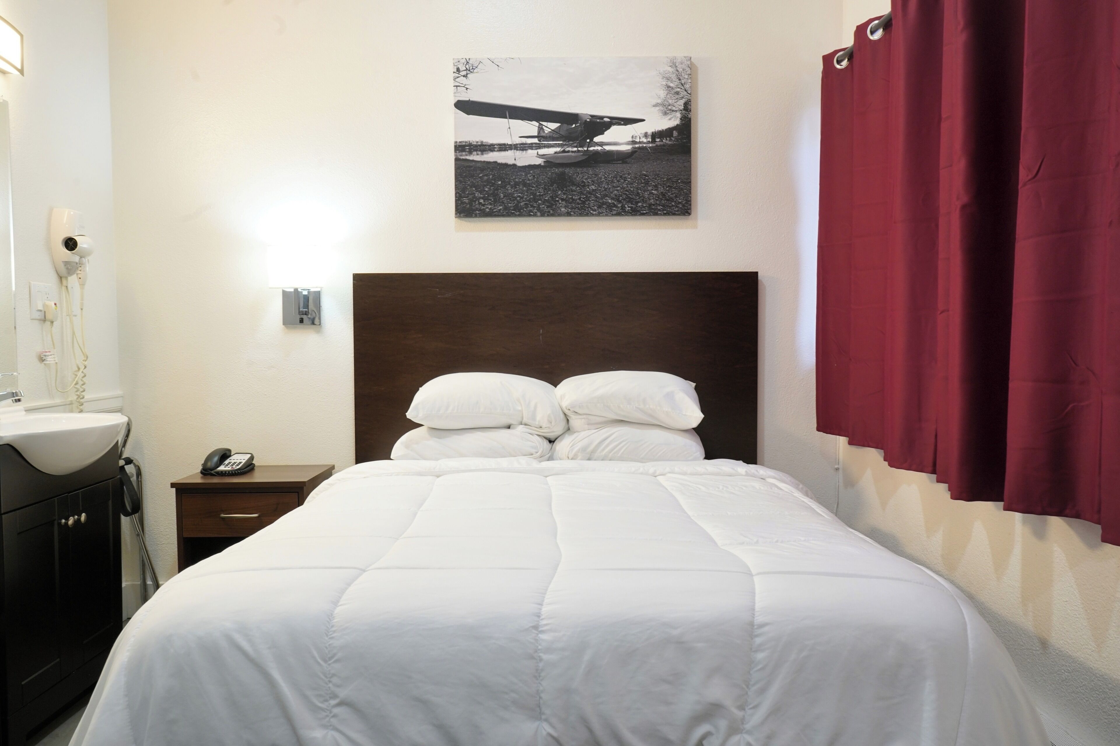 A neatly made bed with white bedding, a wooden headboard, a bedside table with a lamp and a phone, a wall-mounted picture, and a red curtain.