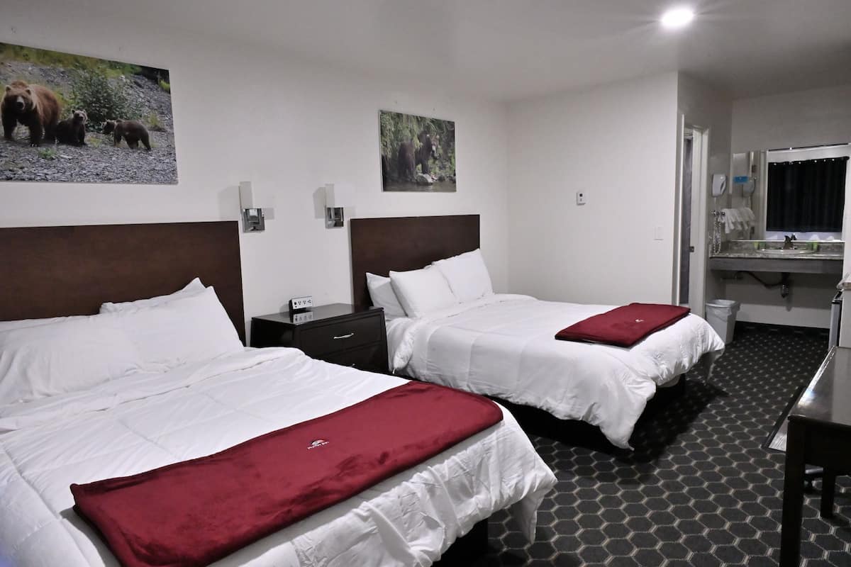 Two beds with white bedding and red throw pillows, a nightstand with a phone, a small desk, and a bathroom with a sink and mirror.