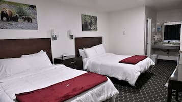 Two beds with white bedding and red throw pillows, a nightstand with a phone, a small desk, and a bathroom with a sink and mirror.