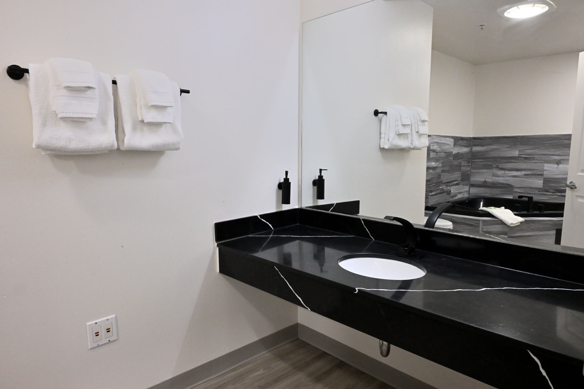Un baño moderno con una encimera negra, un espejo grande y dos toallas blancas colgadas en ganchos.