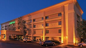 Un hotel de varios pisos con fachada plana, entrada visible y un estacionamiento con automóviles.
