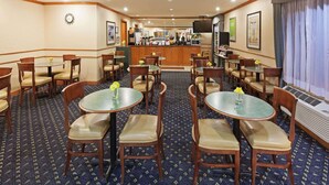 Un comedor de hotel con mesas y sillas redondas, una cocina al fondo y un ventanal con cortinas.