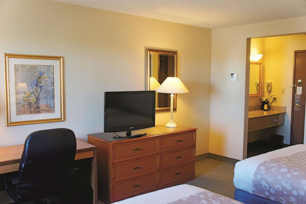 A hotel room with a bed, a desk with a television, a dresser, a lamp, a framed picture on the wall, and a mirror.