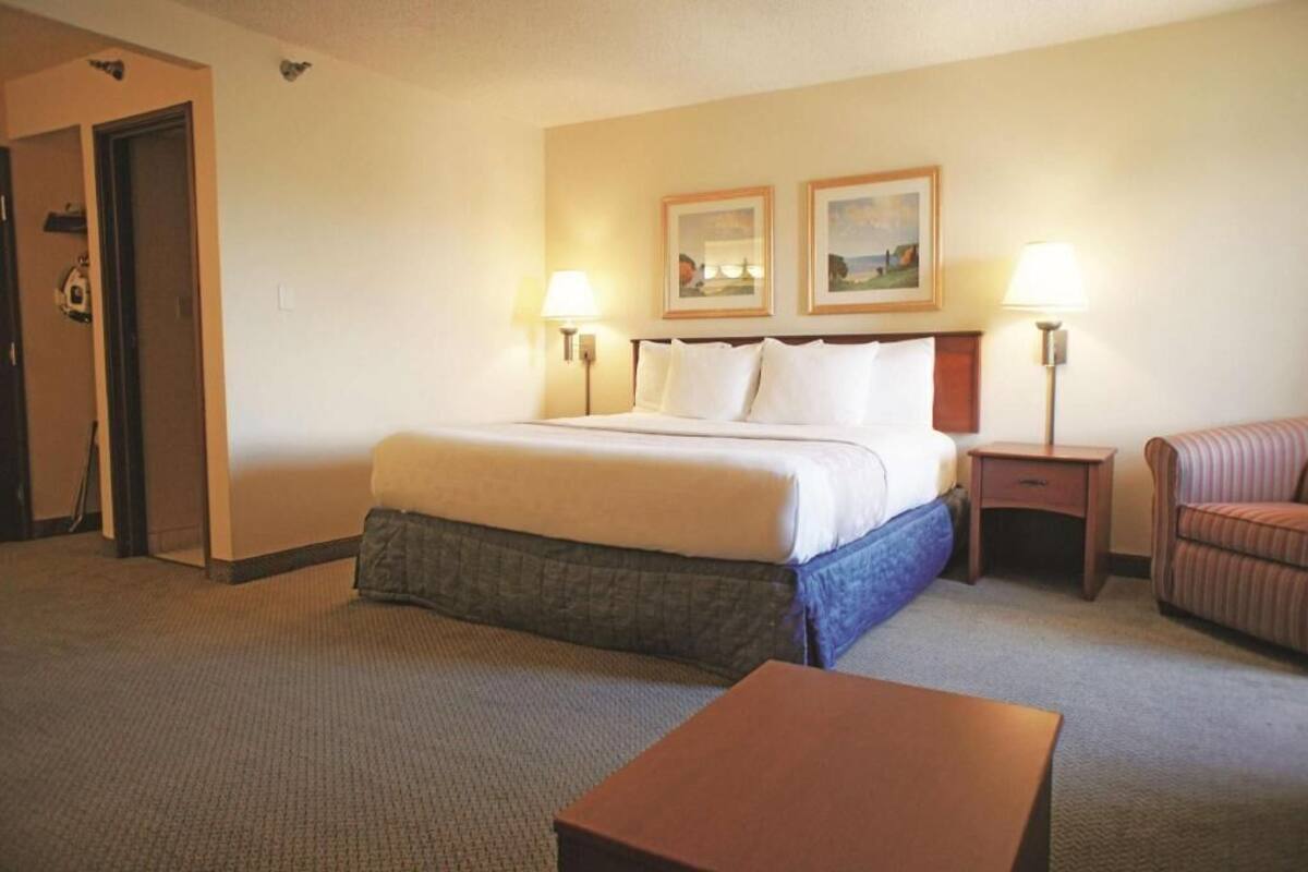 A hotel room with a bed, two bedside lamps, a small table, a chair, and two framed pictures on the wall.