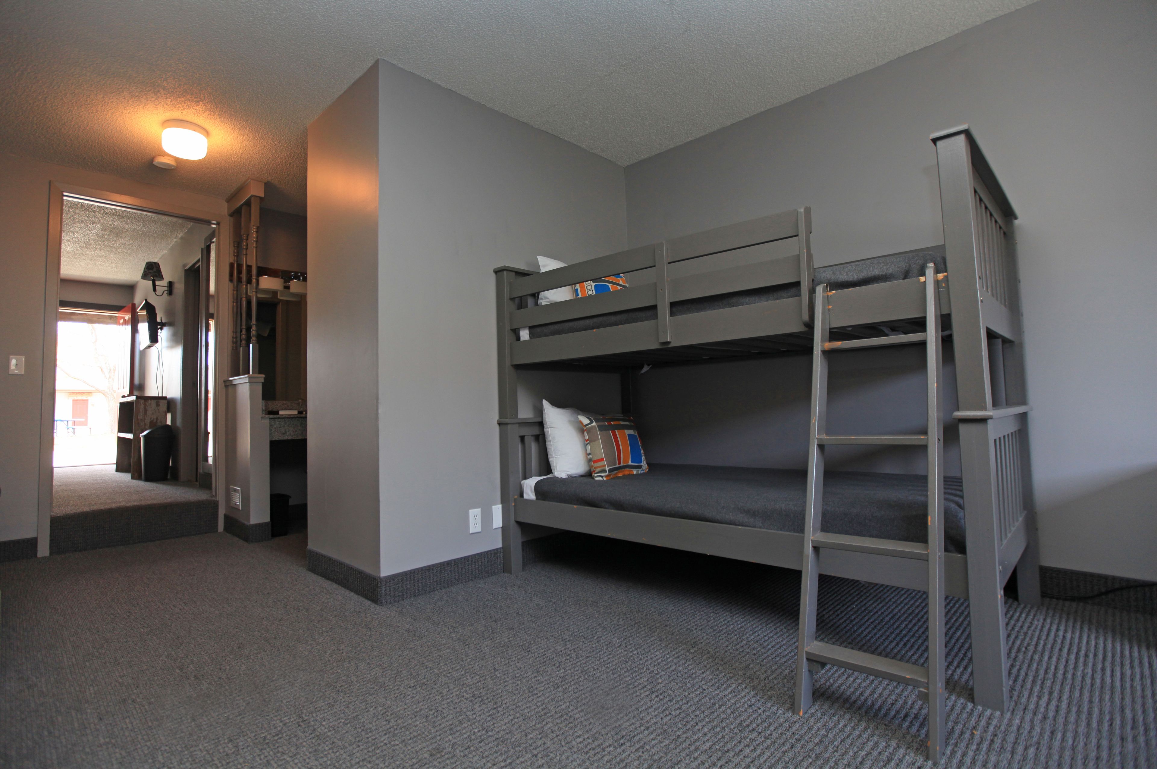 A bunk bed room with a ladder, a door leading to another room, and a carpeted floor.