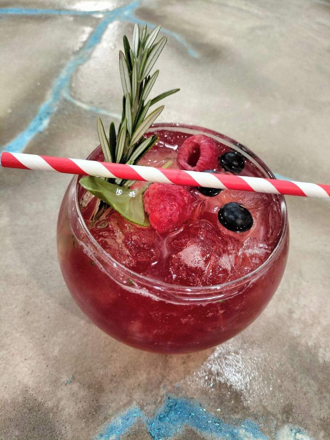 Un verre d’une boisson rouge, servie avec des glaçons, et décorée d’une branche de romarin, de framboises et de mûres, ainsi que d’une paille rayée de rouge et de blanc.