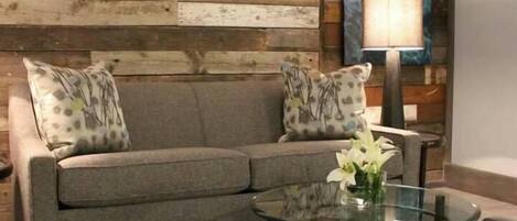 A living room with a grey sofa, patterned pillows, a glass coffee table, and a painting on the wall.