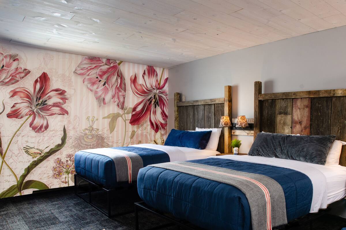 Un dormitorio con dos camas, papel tapiz floral, cabecera de madera y una mesita de noche con lámpara.