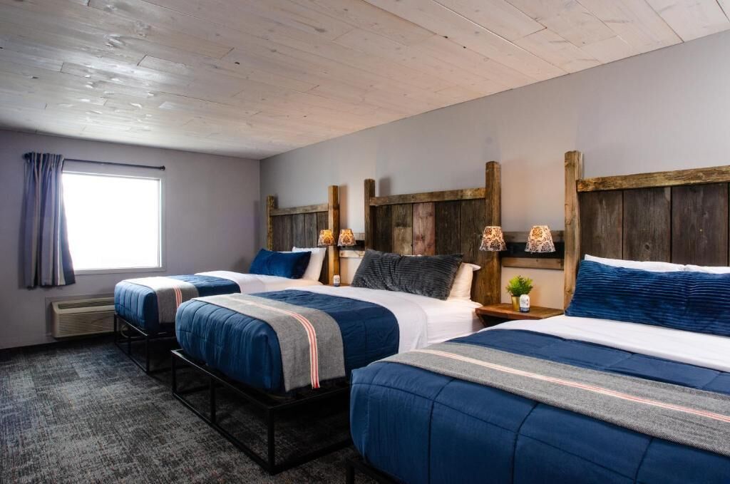 Habitación de hotel con dos camas, una ventana con cortinas, un cabecero de madera y una mesita con lámpara.