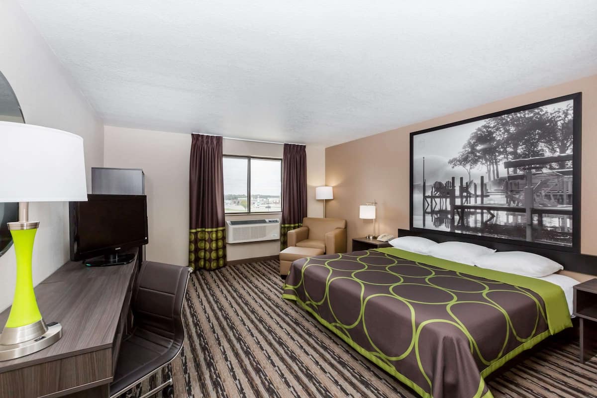 Une chambre d’hôtel avec un lit, un bureau doté d’une lampe, une télévision, une chaise, une fenêtre avec des rideaux et un grand tableau accroché au mur.
