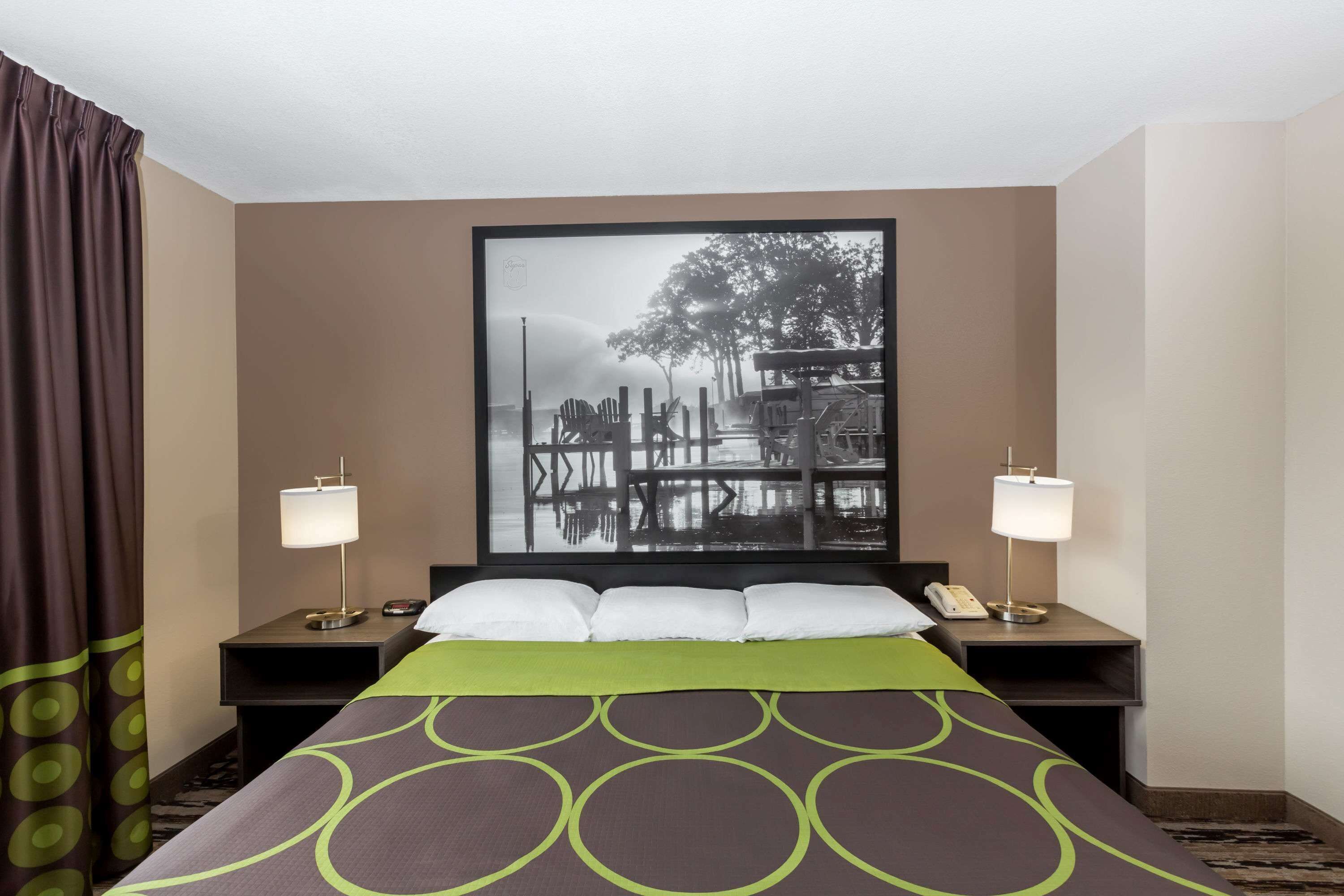 A hotel room with a bed, two bedside tables, a nightstand, a large framed picture on the wall, and a window with curtains.