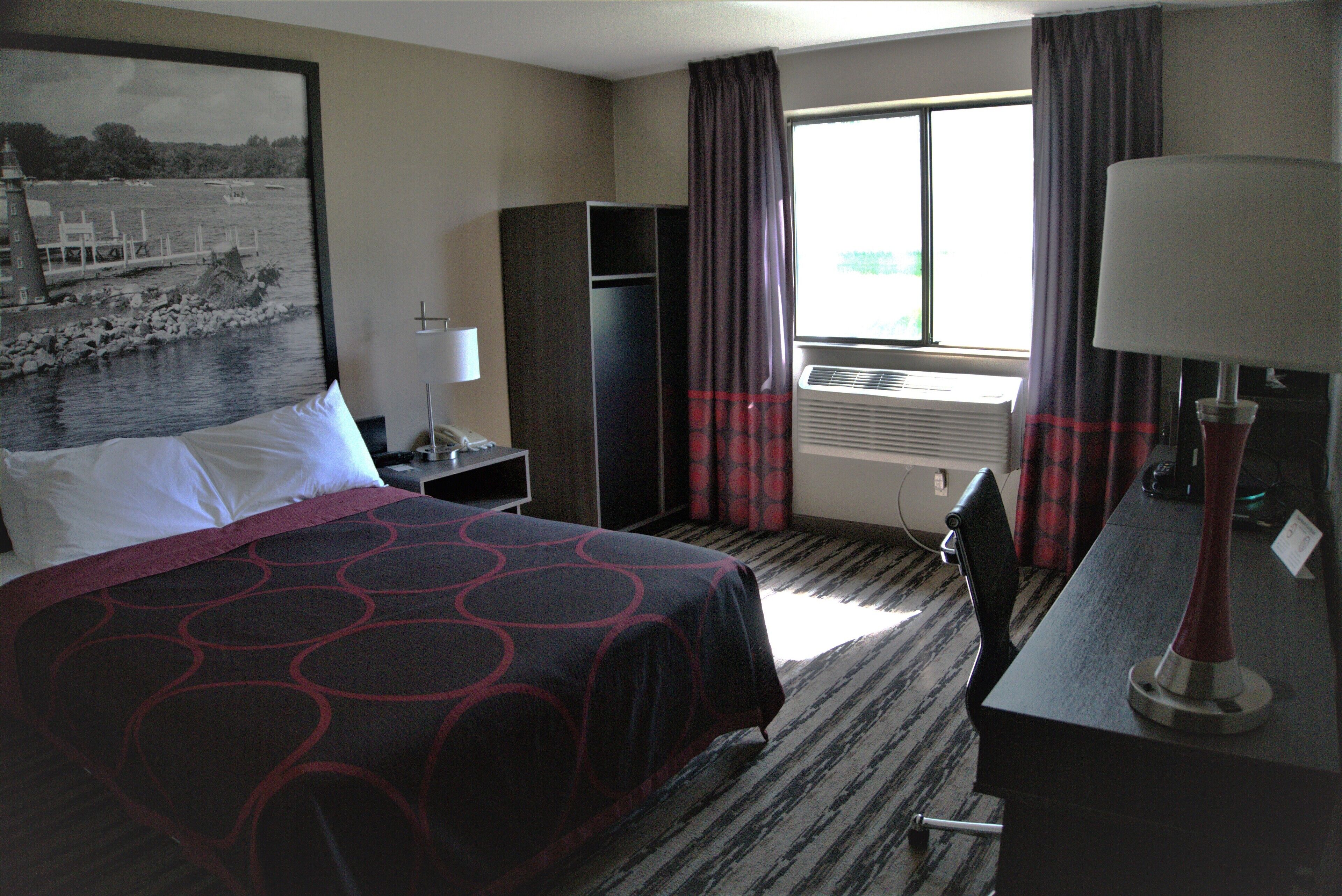 Habitación de hotel con cama, escritorio, silla, lámpara, ventana con cortinas, aire acondicionado y un cuadro en la pared.