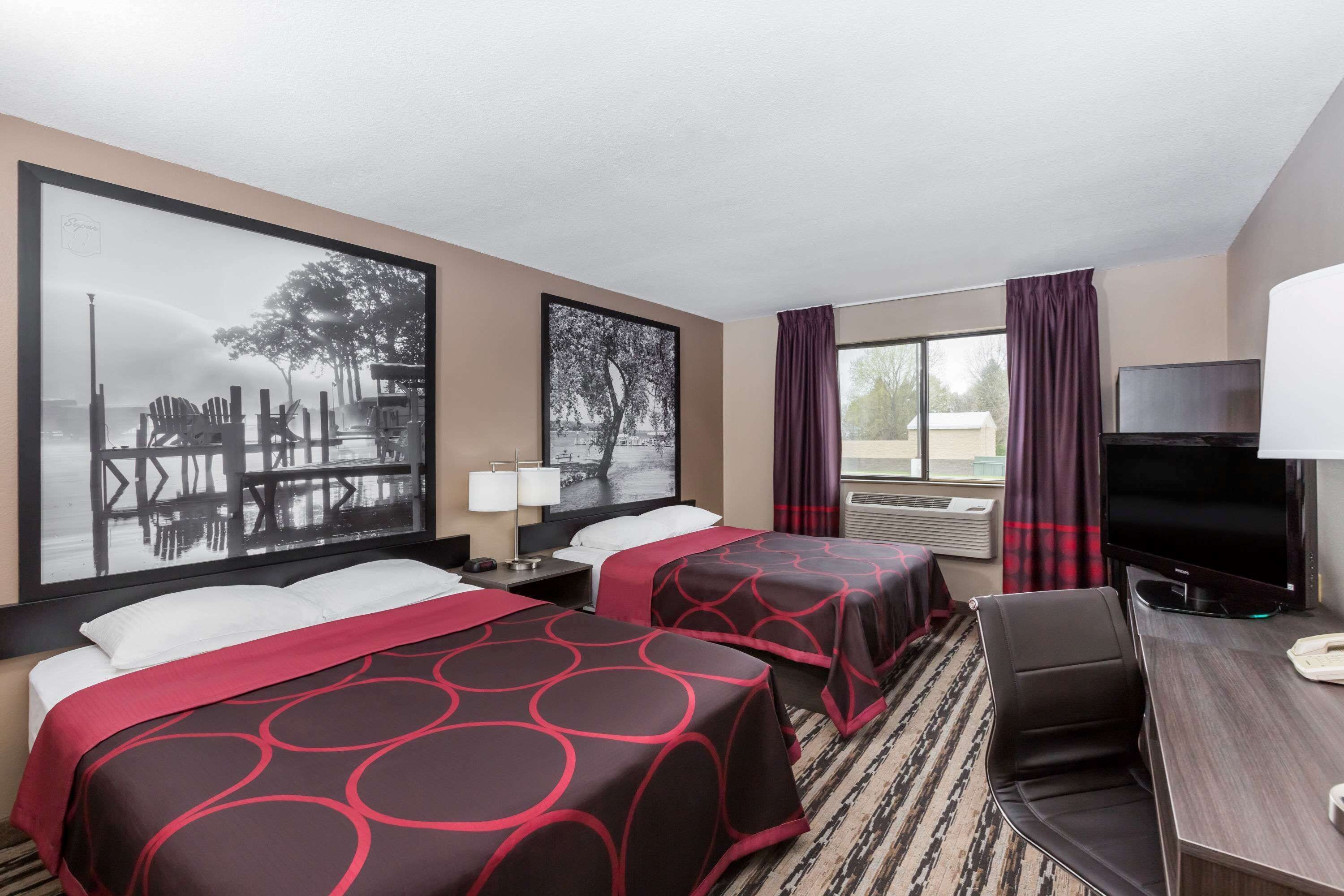 A hotel room with two beds, a desk with a TV, and large windows with curtains.