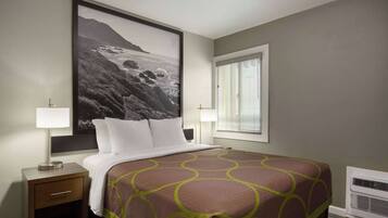 A hotel room with a large bed, a nightstand with a lamp, a window with blinds, and a wall-mounted air conditioner.