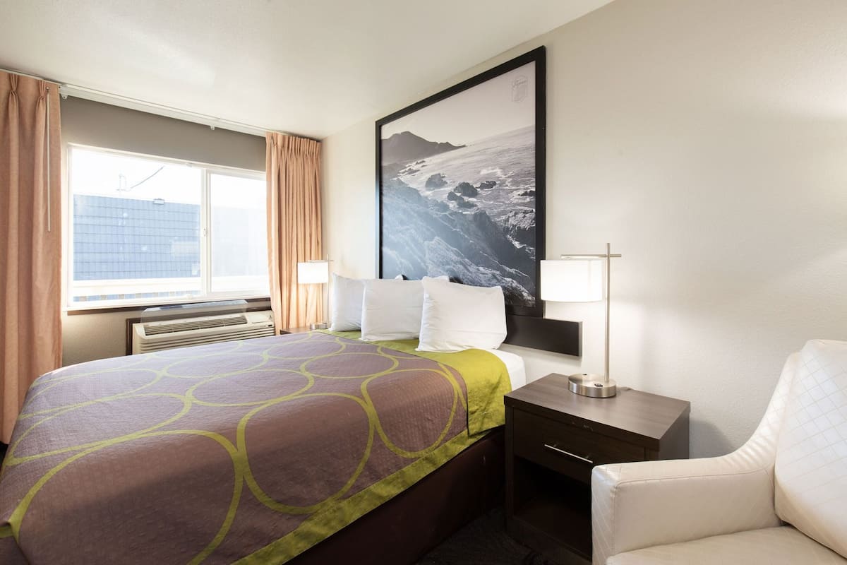 A hotel room with a bed, a nightstand, a lamp, a chair, a window with curtains, and a framed picture on the wall.