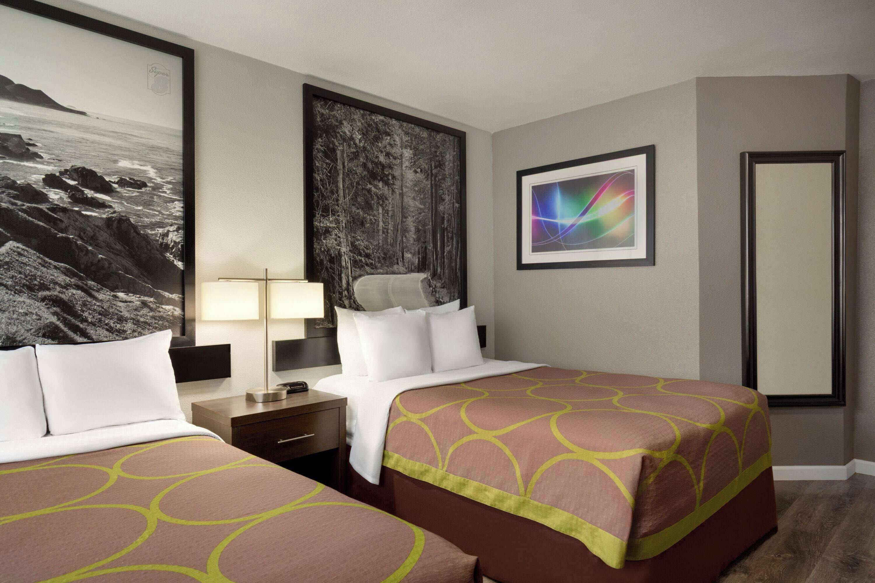 A hotel room with two beds, a nightstand, a lamp, a large framed picture of a forest, and a framed abstract art piece.