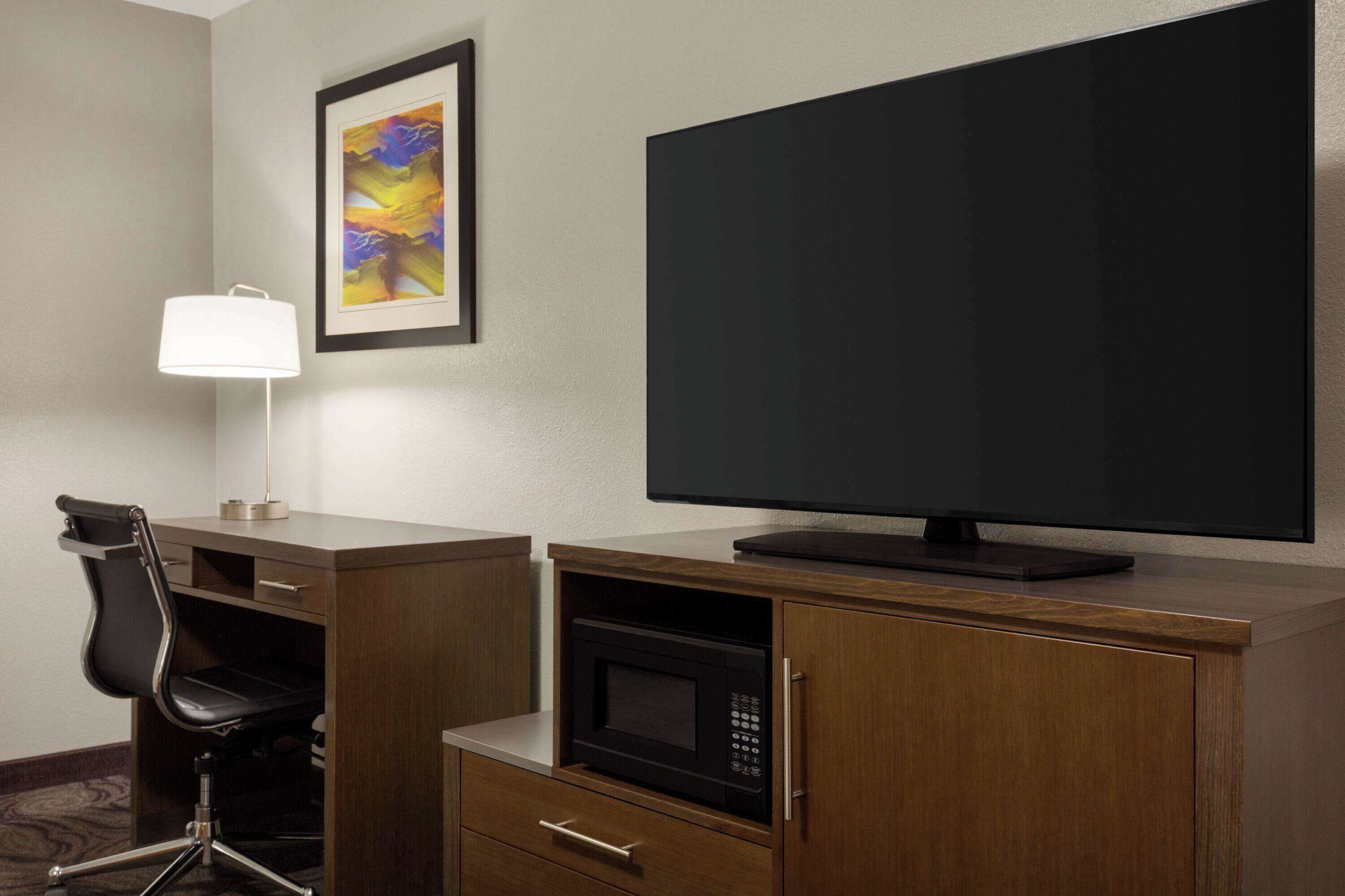 A hotel room with a desk, a chair, a television, a microwave, and a framed picture on the wall.