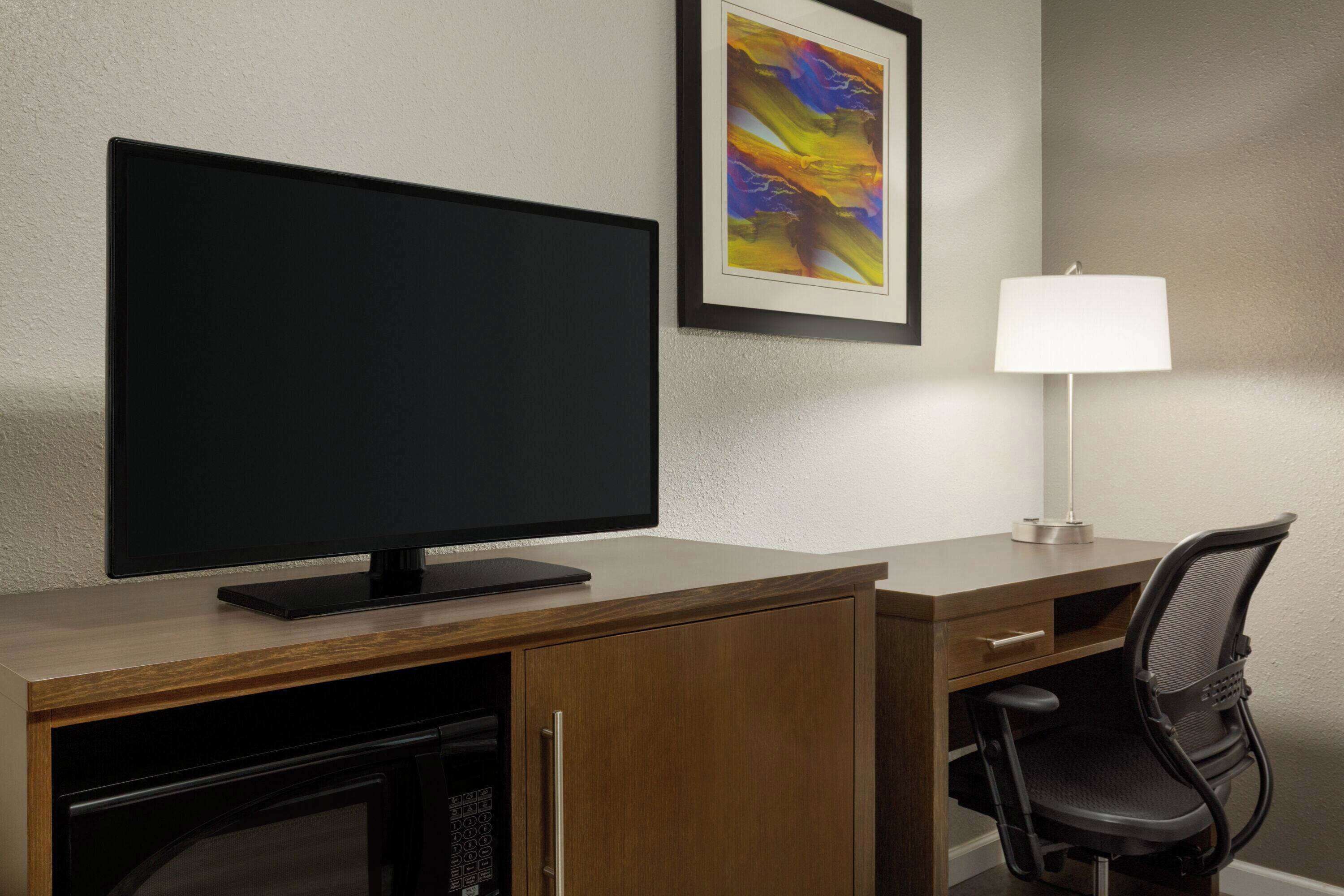 A hotel room with a flat-screen TV, a desk with a lamp, a chair, and a framed abstract painting on the wall.