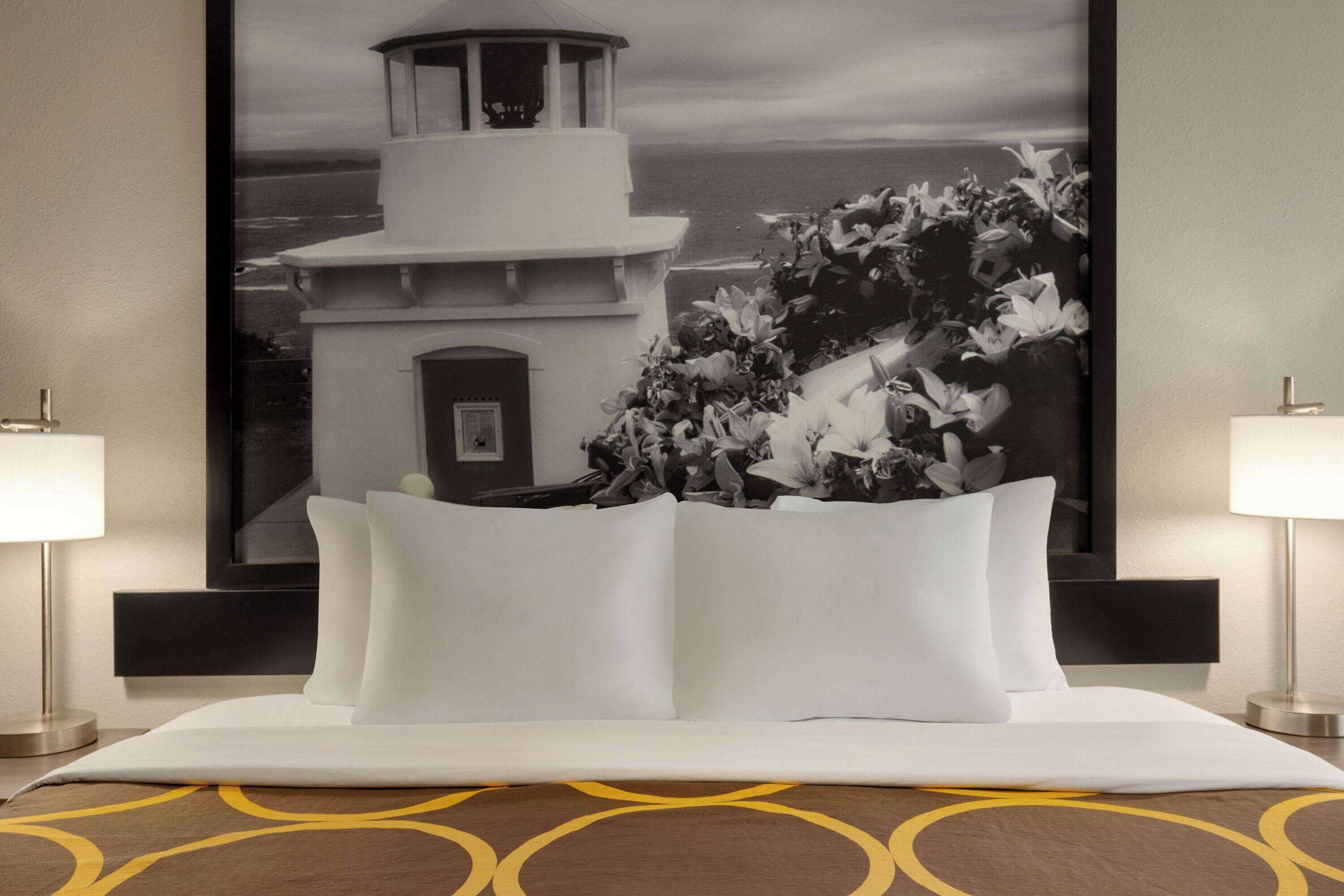 Ein Hotelzimmer mit einem Bett, zwei Nachttischlampen, einem großen gerahmten Bild eines Leuchtturms und einem gemusterten Teppich.
