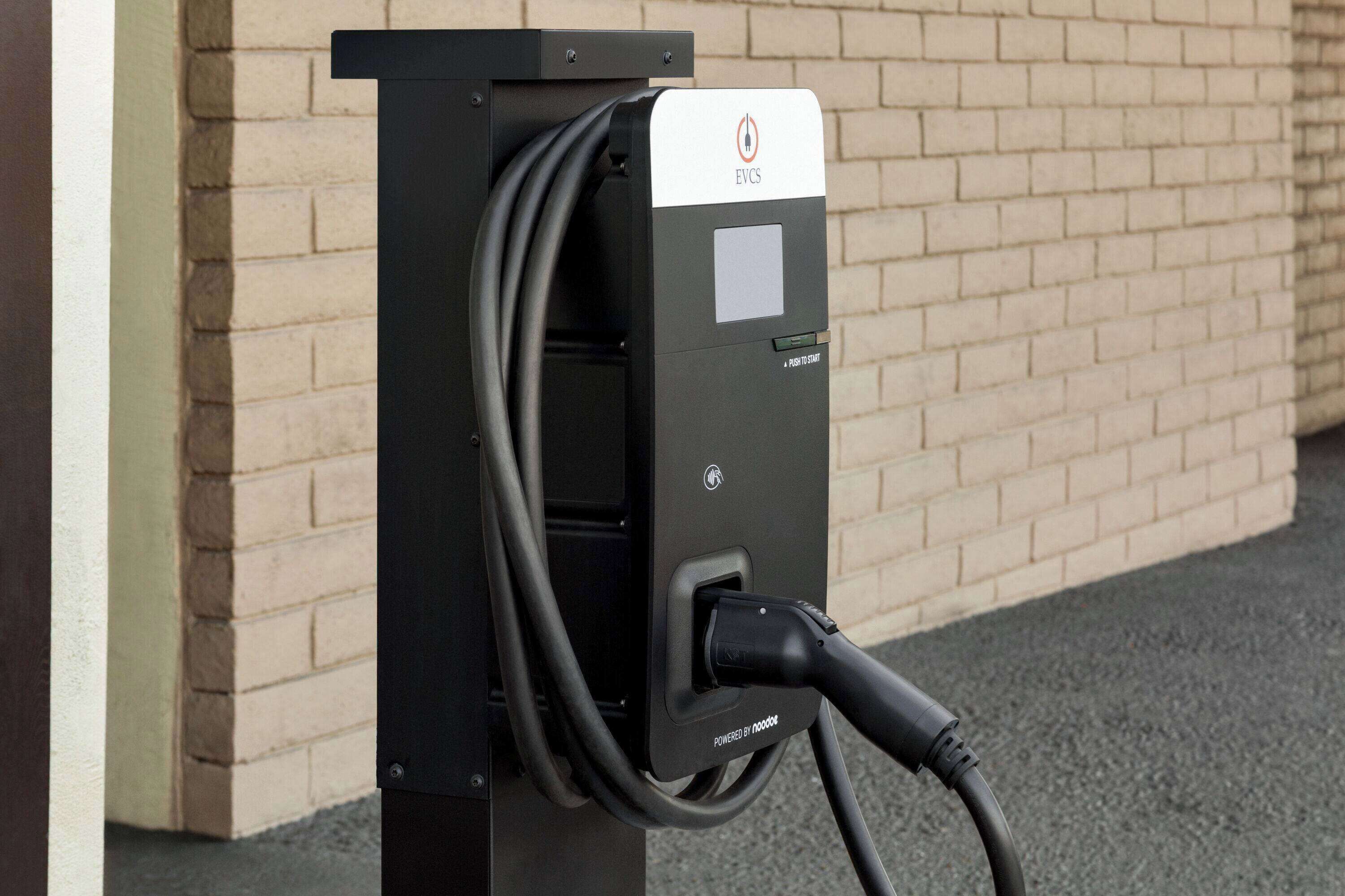 A black EV charging station with a cable plugged in, labeled "EVCS," against a brick wall.