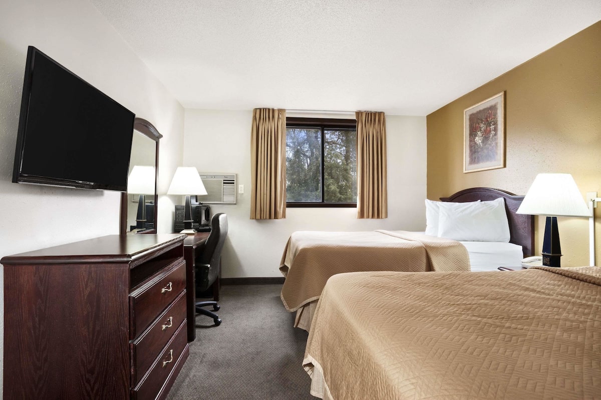 Une chambre d’hôtel avec deux lits, un bureau avec une chaise, une commode, une télévision et une fenêtre avec des rideaux.