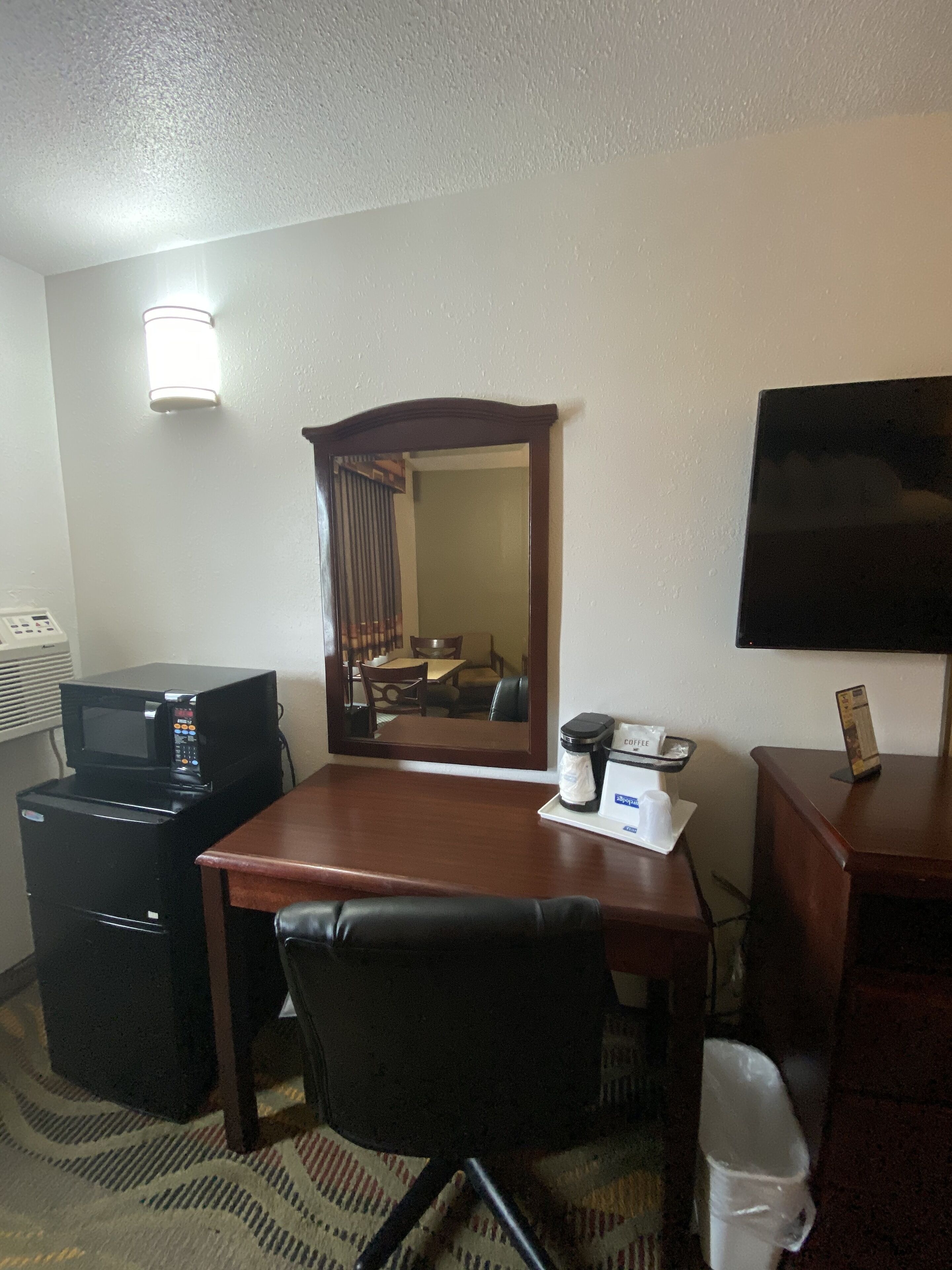 Ein Hotelzimmer mit einem Holztisch, einem schwarzen Bürostuhl, einem Fernseher auf einem Standfuß, einer Mikrowelle, einer Kaffeemaschine und einem Mülleimer.