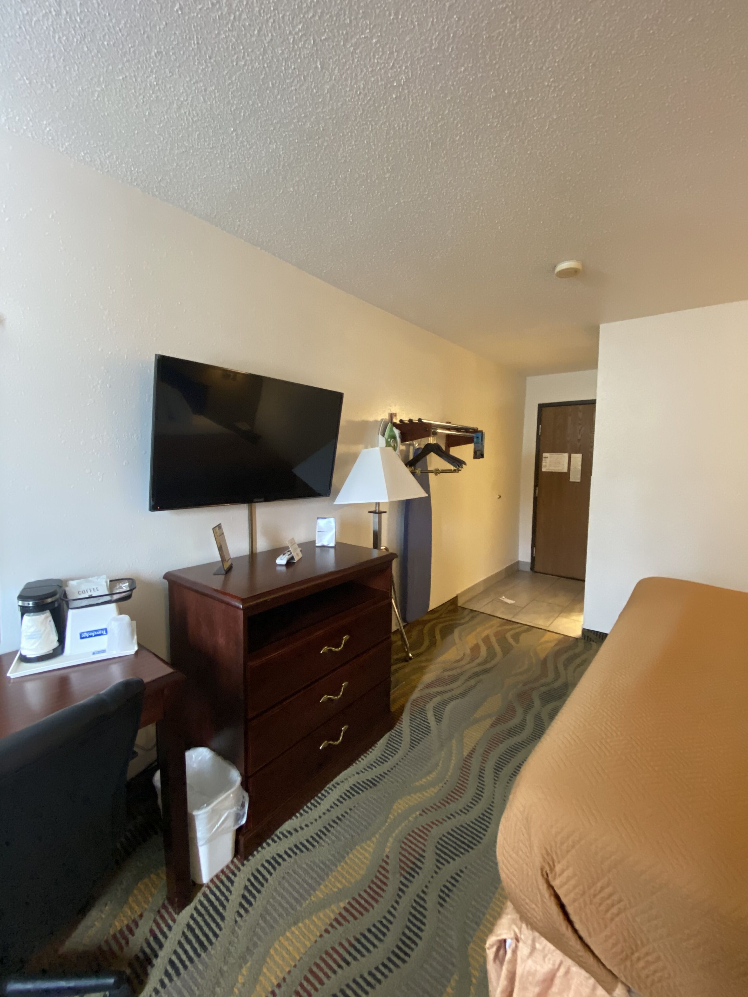 A hotel room with a flat-screen TV mounted on the wall, a wooden desk with a coffee maker, a lamp, and a bed with a patterned bedspread.