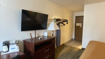 A hotel room with a flat-screen TV mounted on the wall, a wooden desk with a coffee maker, a lamp, and a bed with a patterned bedspread.
