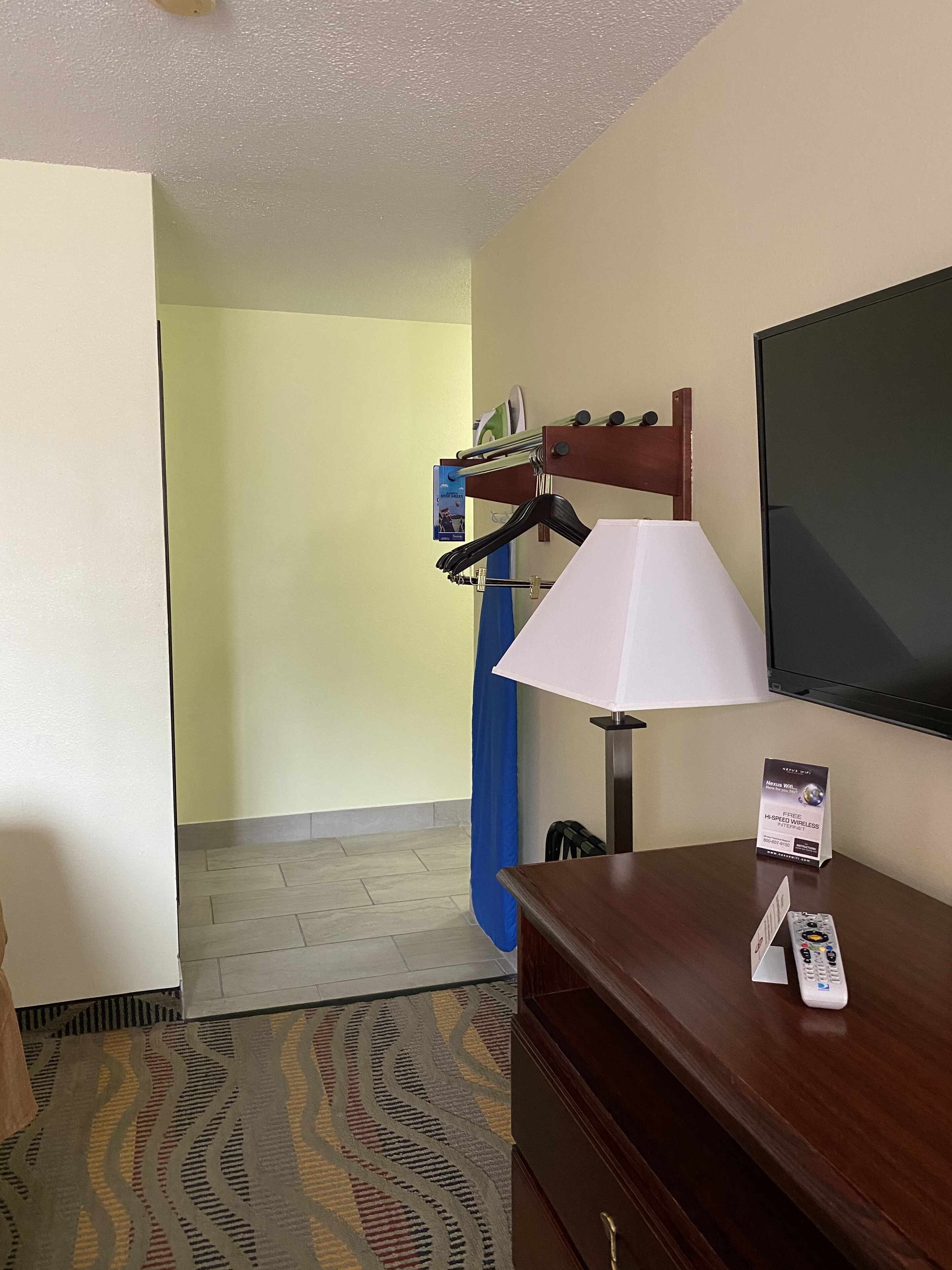 A hotel room with a wooden dresser, a white lamp, a blue curtain, and a television mounted on the wall.