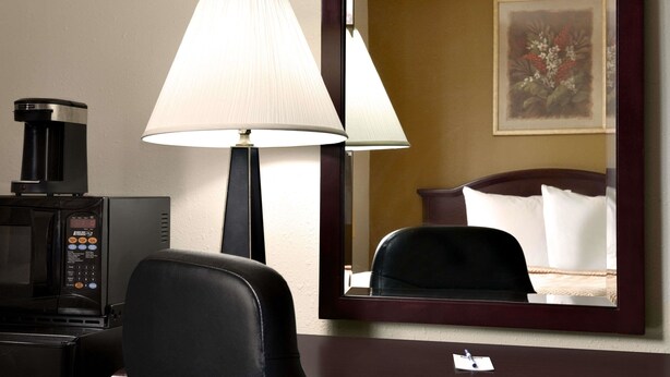 Ein Hotelzimmer mit einem Bett, einem Schreibtisch mit Lampe, einer Kaffeemaschine und einem Spiegel, der das Bett und ein Bild an der Wand reflektiert.