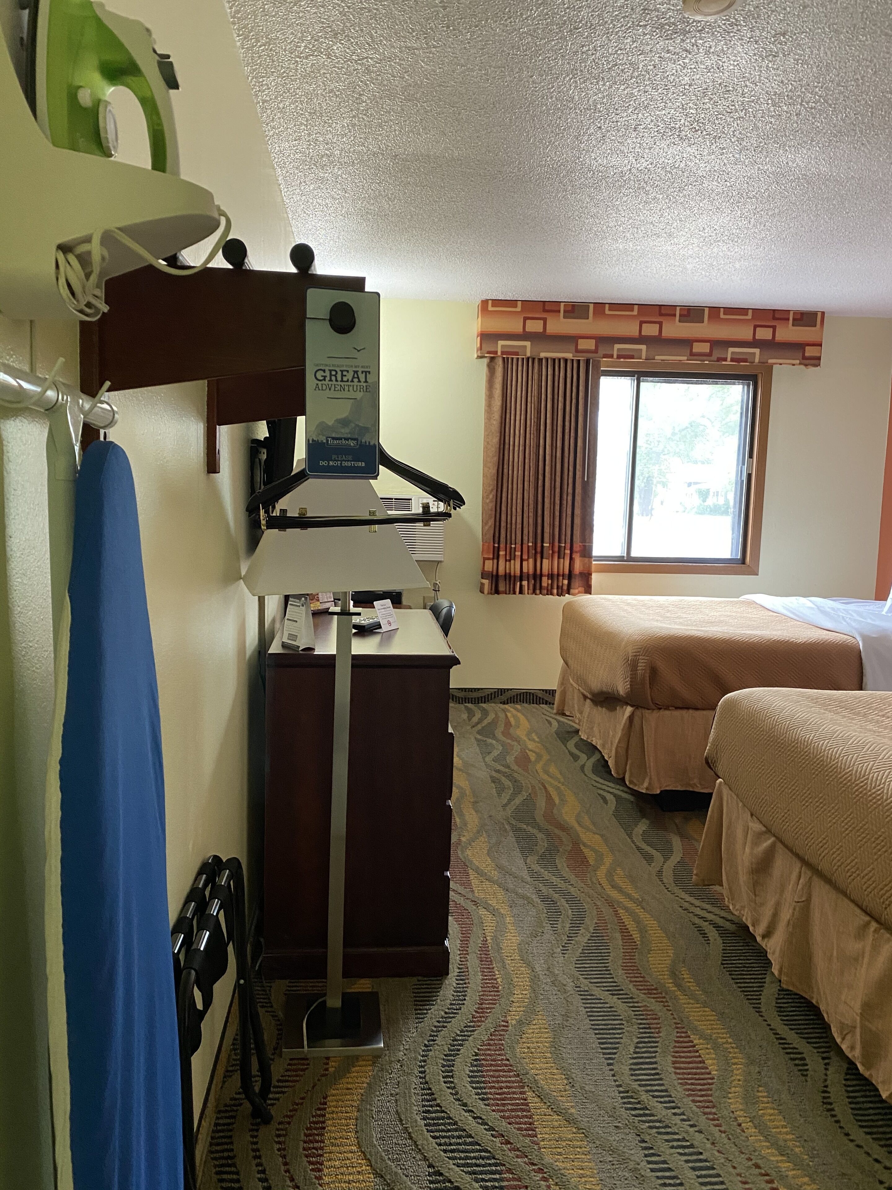 A hotel room with a bed, a desk with a computer, a window with curtains, and a patterned carpet.
