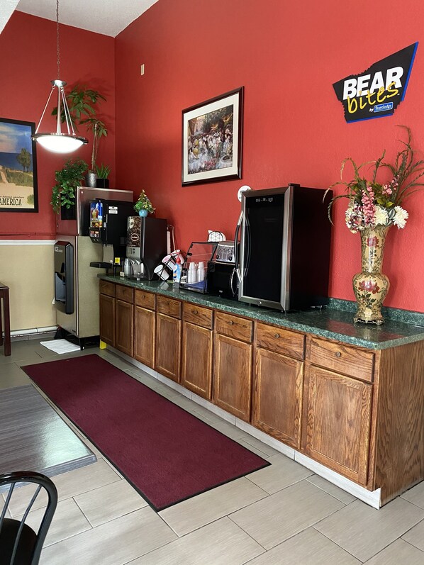 A kitchen with wooden cabinets, a refrigerator, a coffee machine, and a microwave. There is a painting on the wall and a vase with flowers on the counter. - Travelodge by Wyndham La Porte/Michigan City Area (La Porte)
