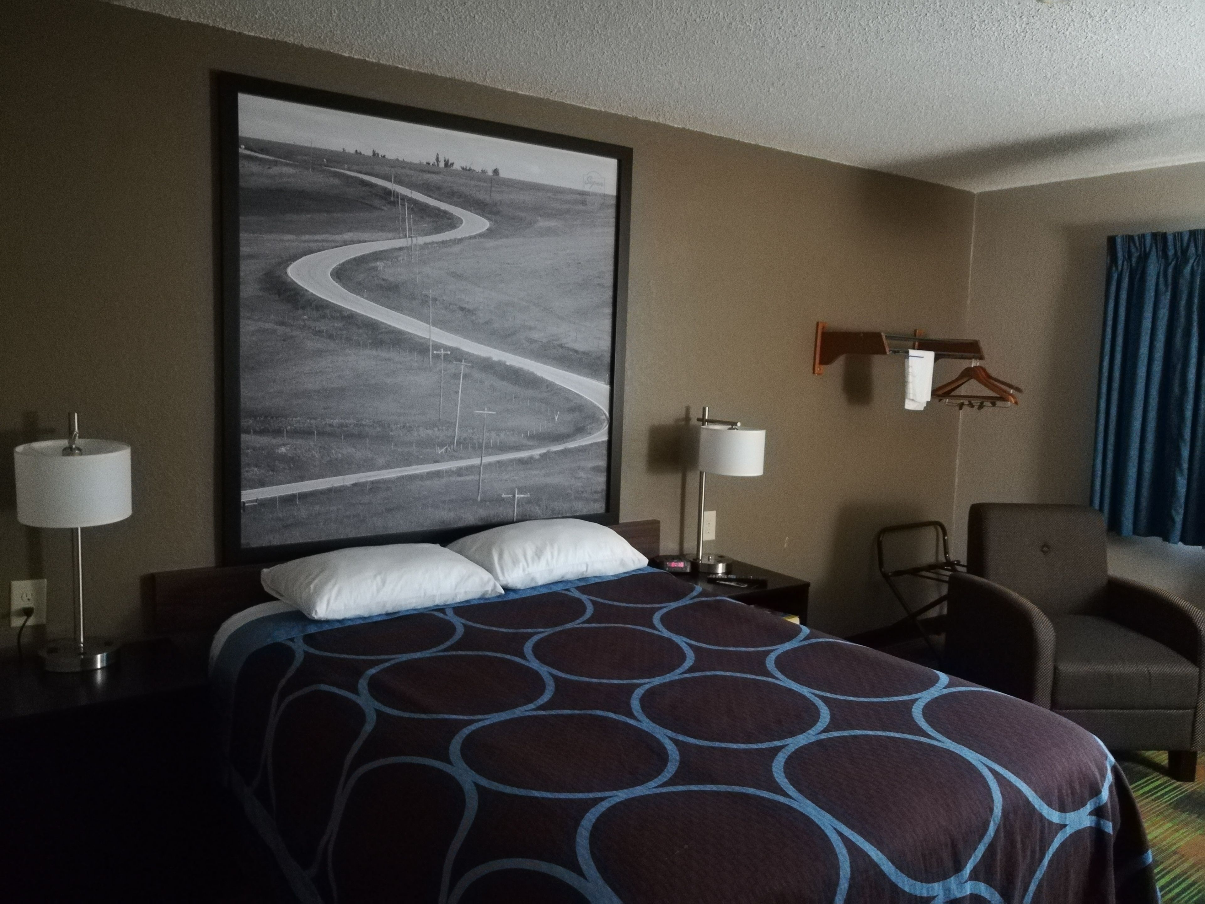 A hotel room with a large bed, two bedside lamps, a chair, a desk, and a large framed picture on the wall.