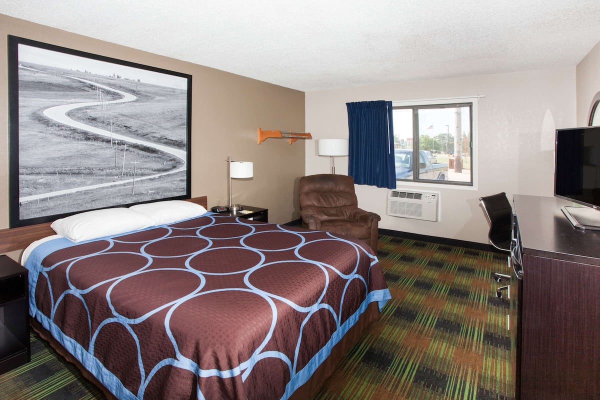 A hotel room with a large bed, a desk with a computer, a chair, a window with curtains, and a wall-mounted TV.