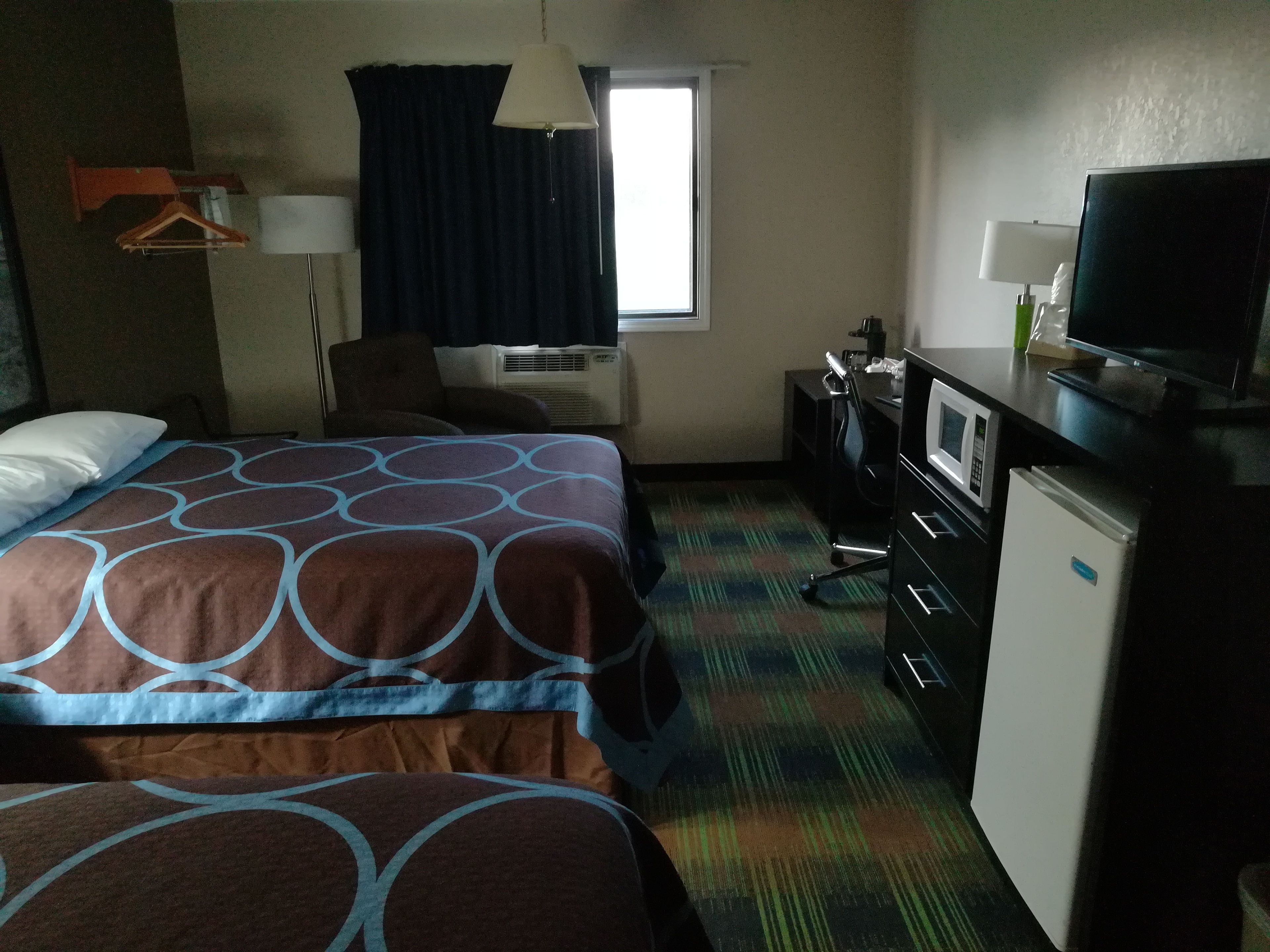 Habitación de hotel con dos camas, un escritorio con televisor, una silla, un refrigerador pequeño y una ventana con cortinas.
